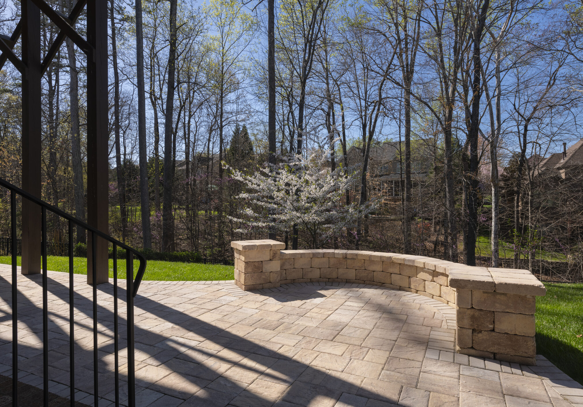 Picturesque backyard view in spring season with patio pavers and stone wall, blooming white cherry tree, and spring colored woods in the background.